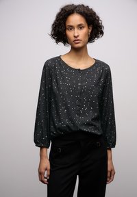 Blusa nera con bottoni, decorata con macchie bianche e dorate, scollo rotondo e maniche lunghe a sbuffo. Abbinata a pantaloni sartoriali neri.