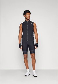 Sportininkas, dėvintis juodą rankovių neturinčią dviračių liemenę, šortus, šalmas, pirštines, baltas kojines ir baltus dviračių batus, stovintis priešais paprastą foną.