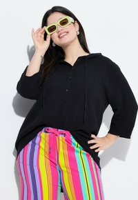 Top nero a maniche lunghe con cappuccio e maniche a tre quarti, abbinato a pantaloni multicolori a righe con strisce rosa, gialle e verdi.