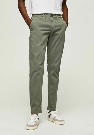 Man wearing olive green slim-fit pants, white sneakers, and a white shirt with hands in pockets, standing against a plain background.