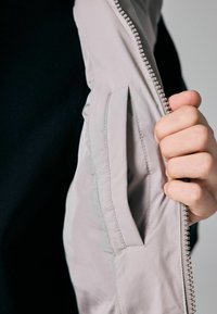 Šviesiai pilkas švarkas su lygia tekstūra, turintis užtrauktuką ir vidinę kišenę, laikomas rankoje prieš juodą foną.