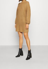 Ljusbrun stickad klänning med hög krage, texturerad mönster och långa ärmar, tillsammans med svarta klackade ankelstövlar.