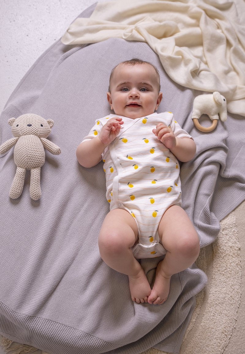 Lichtgrijze gebreide deken met een baby in een witte romper, versierd met gele citroenpatronen; een beige gehaakte berenknuffel en een houten bijtring in de buurt.