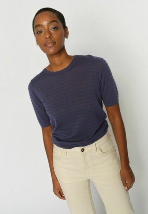 Woman with short hair wearing a textured navy short-sleeve top and light beige pants standing against a plain light background.