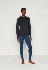 Γυναίκα που στέκεται ξυπόλητη, φορώντας ένα μαύρο μπλουζάκι με μακρύ μανίκι και μικρό λογότυπο, καθώς και εφαρμοστά μπλε κολάν, μπροστά σε μια απλή υπόβαθρο.