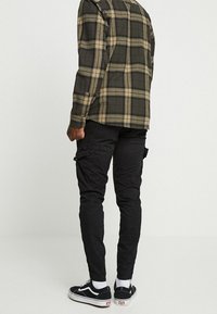 Man i svarta cargo-byxor, svart och beige rutig skjorta, vita strumpor och svarta sneakers som står mot en enfärgad ljus bakgrund.