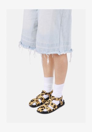 Sandali s potiskom leoparda z usnjem podobno teksturo, črnimi podplati in nastavljivimi trakovi, v kombinaciji z belimi nogavicami do gležnjev in strganimi jeans kratkimi hlačami.