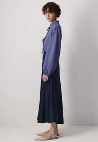 Jupe longue bleu marine associée à un chemisier rayé bleu et blanc avec un col montant et des manches volumineuses, complétée par des ballerines pointues beiges.