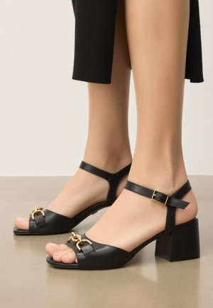 Pieds portant des sandales à talons blocs en cuir noir, à bout ouvert, avec des brides à la cheville et des ornements en chaîne dorée sur un sol lisse.