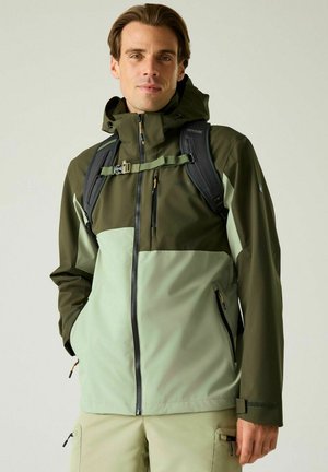 Young man wearing two-tone green outdoor jacket and black backpack, standing with hands in jacket pockets against plain background.