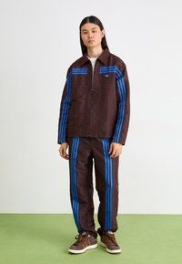 Brun mönstrad jacka och byxor med blå ränder, utrustade med dragkedja framtill och avslappnad design. Komplett med bruna sneakers.