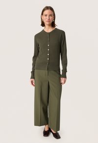 Olijfgroene geribde cardigan met lange mouwen en knoopsluiting, gecombineerd met olijfgroene wijde broek en donkerbruine schoenen.
