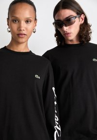 Schwarze Langarm-T-Shirts mit gestickten grünen Logos auf der Brust und weißen Schriftzügen an den Ärmeln. Der Stoff wirkt glatt und dehnbar.