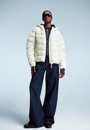 Crème kleurige pufferjack met capuchon en ritsluiting, gecombineerd met wijde blauwe denim broek. Zwarte zonnebril en gelaagde top zichtbaar.