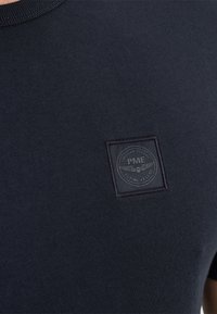 Camiseta de manga corta azul marino con un escote redondo y una tela texturizada. Presenta un parche cuadrado con el logotipo "PME" en relieve en el pecho.