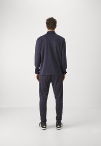 Persoon die met de rug naar de camera staat, draagt een donkerblauw trainingspak en zwarte Adidas sneakers op een effen lichte achtergrond.