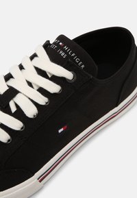 Schwarze Leinensneaker mit weißen Schnürsenkeln, ausgestattet mit einer gemusterten Textur, einem kleinen Logo in Rot, Weiß und Blau sowie einer kontrastierenden gestreiften Sohle.