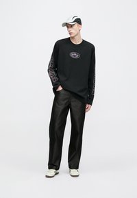 Juoda ilgomis rankovėmis marškinėliai su purpuriniu logotipu ant krūtinės ir rankovių, derinami su laisvai krentančiomis tamsiomis kelnėmis ir baltomis sportinėmis bateliais. Ant galvos balta kepurė.