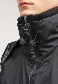 Czarna, wodoodporna kurtka z wysokim kołnierzem, teksturowaną tkaniną, regulowanymi stoperami i wyraźnym logo z przodu.