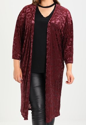 Vrouw die een lange bordeauxrode fluwelen cardigan met bloemmotief draagt over een zwarte top en zwarte leren broek, staand met ontspannen armen langs haar zij.