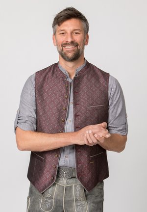 Mann lächelt, trägt traditionelle bayerische Tracht mit gemusterter Weste, kariertem Hemd und bestickter Lederhose mit Hirschgürtelschnalle.