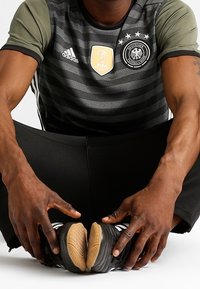 Černé a šedé pruhované sportovní tričko s krátkými rukávy, nese loga FIFA a německého národního týmu, nošeno s černými kalhotami a kopačkami.