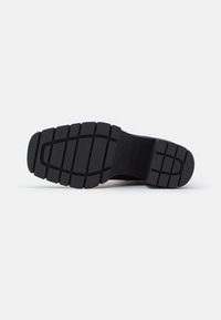Semelle de chaussure noire avec des motifs de crampons profonds conçus pour l'adhérence, présentant des rainures rectangulaires et courbes pour la traction sur diverses surfaces.