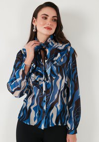 Long-sleeve blouse in sheer fabric featuring a dynamic blue, brown, and cream wave pattern, with ruffled collar and black tie detail.