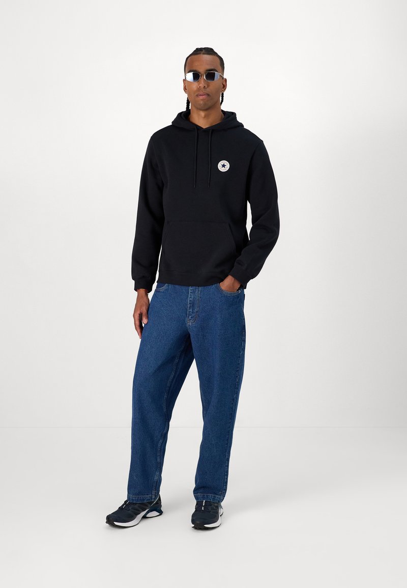 Sudadera de algodón negra con un bolsillo delantero y un logo, combinada con jeans de denim azul de corte holgado y zapatillas negras con detalles en blanco.