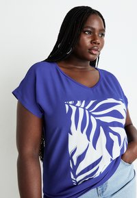 V-neck purple top made of soft fabric, featuring a white leaf pattern on one side. Short sleeves, relaxed fit, paired with light denim.
