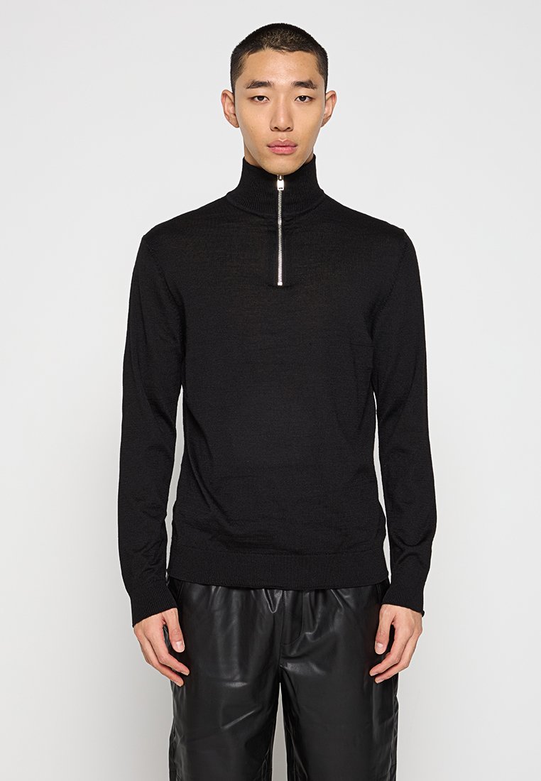 Pull à col zippé noir en maille fine, avec un col montant et un ourlet côtelé. Associé à un pantalon noir ample, créant une silhouette élégante.