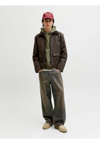 Giacca marrone con cerniera, felpa verde con cappuccio sotto, jeans grigi larghi e calzature beige. Completa il look un cappellino rosso con dettagli del logo.