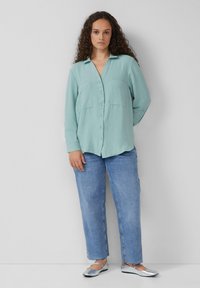 Hellgrünes Hemd mit zwei vorderen Taschen, strukturierter Stoff, kombiniert mit lockeren blauen Jeans und silbernen flachen Schuhen.