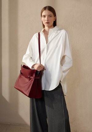 Camicia bianca oversize, pantaloni larghi grigi e una borsa in pelle bordeaux con tracolla e dettagli in metallo, su uno sfondo neutro.