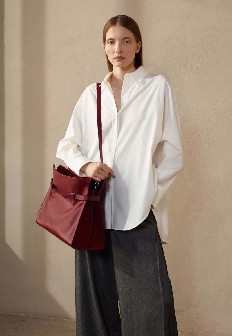 Camisa blanca de gran tamaño, pantalones anchos gris, y un bolso de cuero burdeos con correa y herrajes de metal, todo ello sobre un fondo neutro.