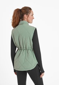 Quilted green vest, lightweight fabric, elastic waistband, sleeveless design, back view showing diamond pattern, worn over a black long-sleeve shirt.