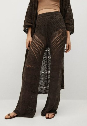 Mujer que lleva pantalones anchos de punto marrón oscuro transparente con patrones geométricos, combinados con un cárdigan largo a juego y sandalias marrones.