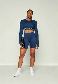 Woman in navy blue long-sleeve crop top and matching bike shorts holding black water bottle, standing against plain light background.