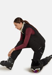 Gilet nero imbuccato sopra una camicia a maniche lunghe bordeaux, pantaloni da sci neri e scarponi da sci. Gli accessori includono una sciarpa crema e occhiali da sci specchiati.