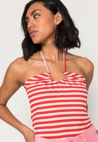 Top halter riscado em vermelho e branco com detalhe franzido no decote, feito de tecido canelado. A modelo está a usá-lo com calções rosa claro.