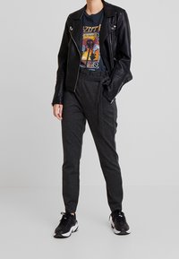 Osoba v černé kožené bundě, tričku s grafikou Led Zeppelin, tmavě šedých kalhotách svázaných v pase a černých teniskách stojící proti šedému pozadí.