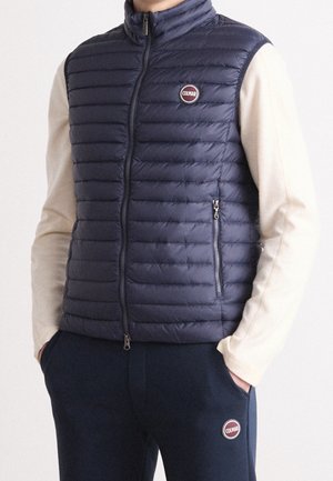 Persona vestida con un chaleco acolchado azul marino con el logo de "Colmar" sobre una camiseta de manga larga beige y pantalones azul marino con el logo a juego, con las manos en los bolsillos.