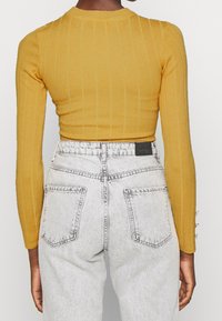 Hořčicový žebrovaný top s dlouhým rukávem v kombinaci se světle šedými džínami s vysokým pasem, které mají dvě zadní kapsy a jemný opotřebovaný vzhled.