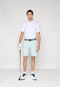 Biela polokošeľa s malým logom, mäta zelené šortky, sivý opasok, biele golfové topánky s modrými detailmi a golfová palica v ruke.