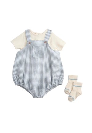 Cremefarbenes Kurzarmhemd unter einem blau-weiß karierten Overall mit Knöpfen und Tasche vorne, kombiniert mit cremefarbenen Socken mit hellblauen Streifen.