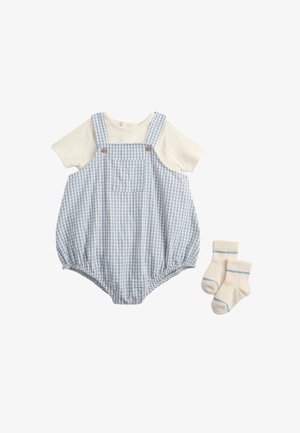 Cremefarbenes Kurzarmhemd unter einem blau-weiß karierten Overall mit Knöpfen und Tasche vorne, kombiniert mit cremefarbenen Socken mit hellblauen Streifen.