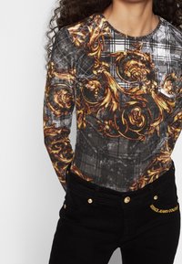 Svart långärmad topp med ett grått rutmönster och ett guldornamenterat mönster. Svarta denimjeans med guldknappar och broderi av "VERSACE JEANS COUTURE".