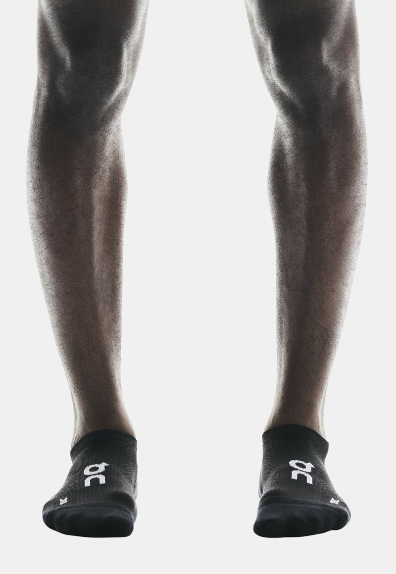 Chaussettes noires de cheville avec logo blanc, offrant un ajustement serré et des poignets côtelés, présentées sur des jambes bien définies sur fond blanc.