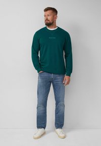 Camicia verde a maniche lunghe con scollatura rotonda, dotata di un piccolo logo sul petto, abbinata a jeans blu e sneaker bianche.