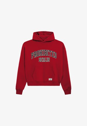 Mann trägt einen roten Hoodie mit dem Text "PROHIBITED BERLIN" und eine schwarze Mütze und schaut nach links vor einem schlichten grauen Hintergrund.